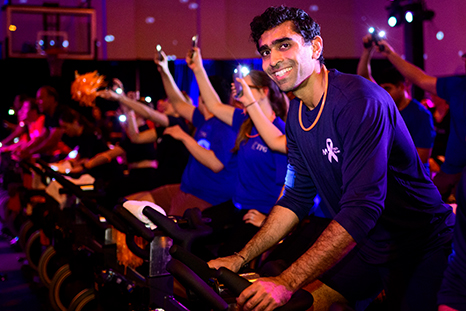 People ride stationary bikes under colorful lights and hold up phones at a Cycle for Survival indoor cycling event. 