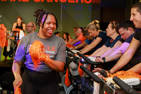 A participant holding orange pom-poms stands next to people on stationary bikes at a Cycle for Survival event. 