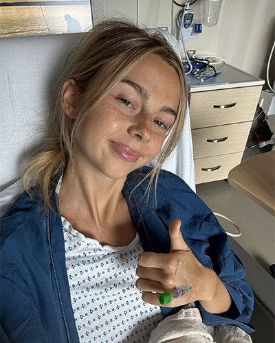 MSK patient Sydney Towle, wearing a hospital gown and smiling, gives a thumbs-up from her bed.