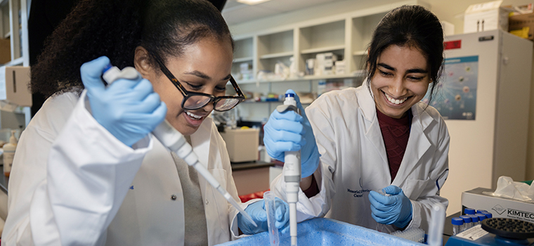 Two research scientists smile in a laboratory while working with pipettes.