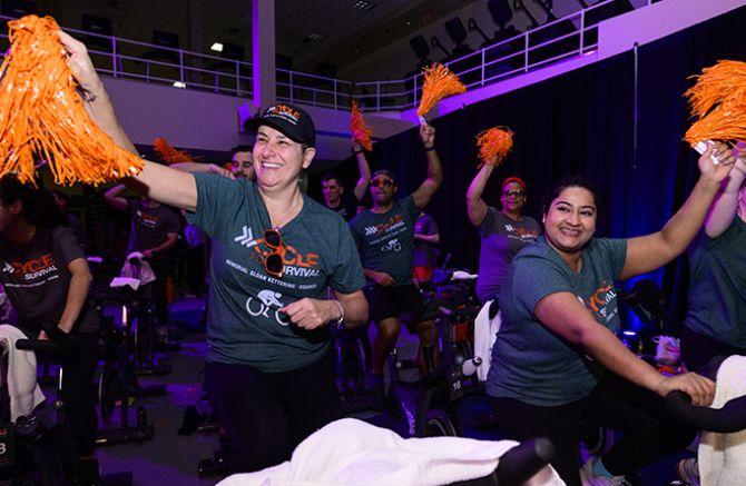 People riding stationary bikes at Cycle for Survival, wearing gray shirts and waving orange pom-poms in a dimly lit event space. 