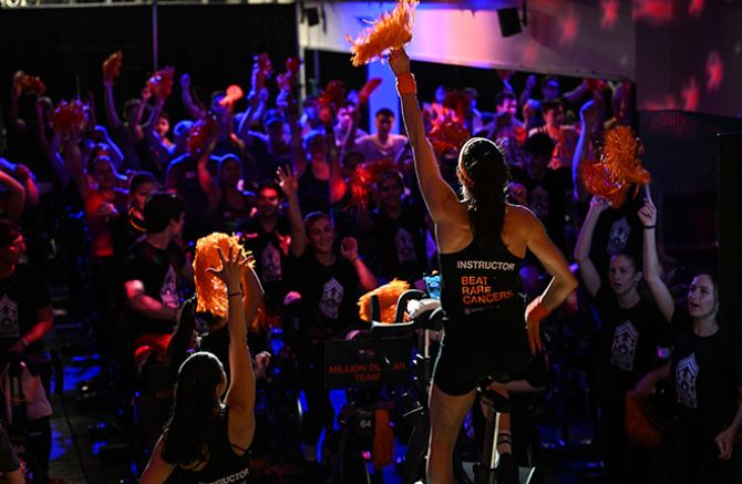 Cycle for Survival indoor cycling event with energetic participants and instructors raising orange pom-poms to support rare cancer research. 