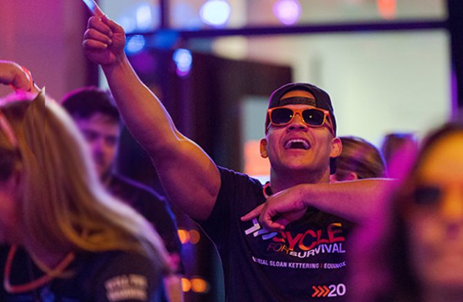 Person enthusiastically pointing upward at a Cycle for Survival event, wearing a branded event t-shirt. 
