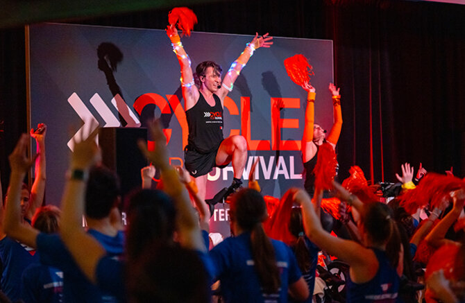 Equinox instructor with orange pom-poms leads a cheering crowd at a Cycle for Survival event.