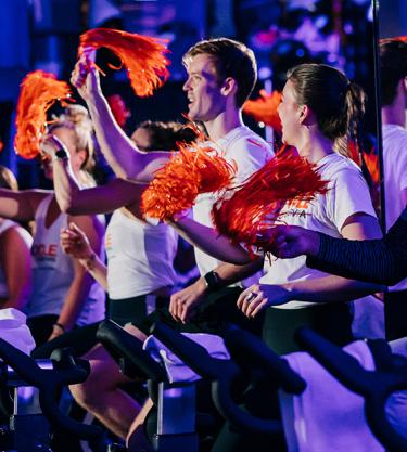 A group of people ride stationary bikes at a Cycle for Survival event. They are smiling and waving orange pom poms, excited to make an impact on rare cancer research at MSK.