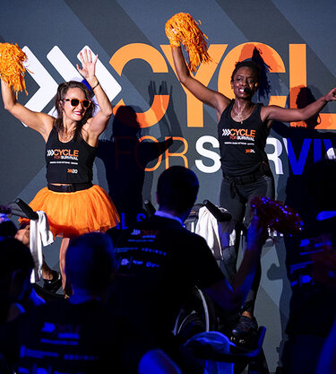 Cycle for Survival instructors lead riders from the stage as participants pedal and wave orange pom-poms during an indoor event.