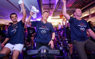 Three men sit on stationary bikes, wearing shirts that say “Team JP Morgan Chase,” each with one hand raised in the air and smiling.