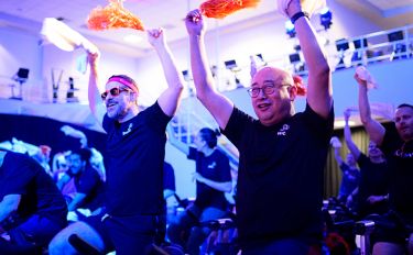 Participants sit on stationary bikes, smiling and raising pom-poms and towels, at a Cycle for Survival event.