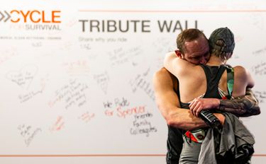 Two participants at a Cycle for Survival rare cancer fundraiser hug in front of a Tribute Wall featuring emotional written messages.
