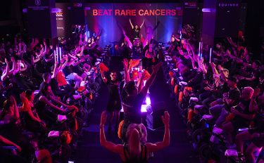Participants at an indoor cycling fundraiser raise their hands beneath a “Beat Rare Cancers” sign in colorful lighting.