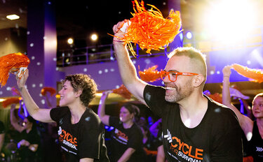 Participants wearing Cycle for Survival shirts cheer with orange pom-poms during an indoor fundraising ride.