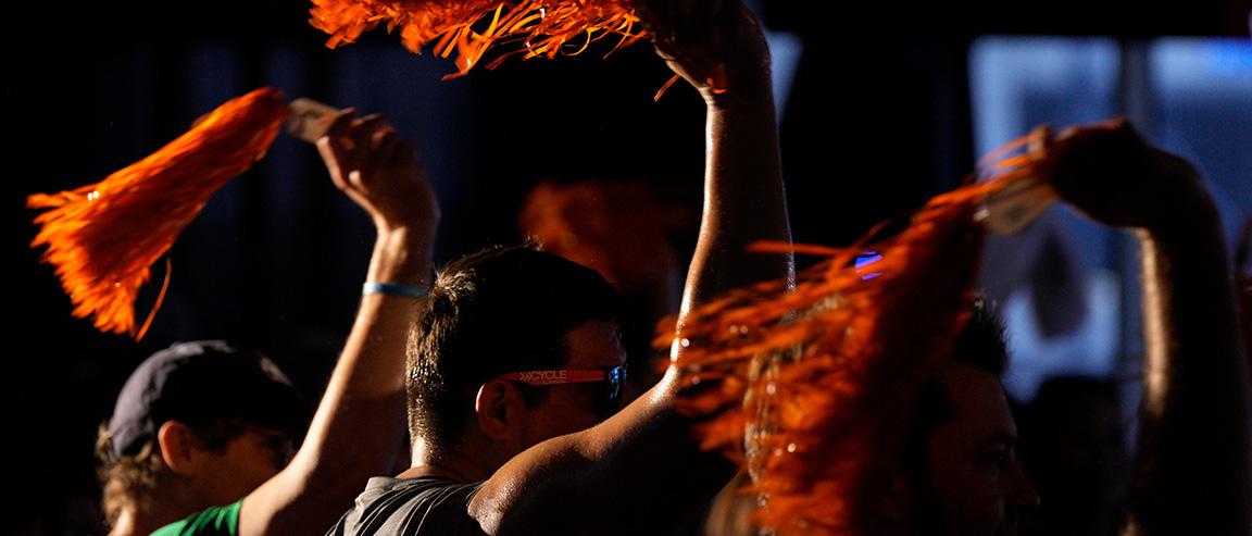 Three people seen from the head and shoulders up, waving orange pom-poms over their heads in a dark room to support a rare cancer fundraiser 