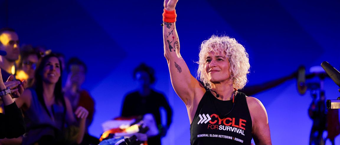 A person with curly blonde hair raises their arm at a Cycle for Survival event, wearing a black tank top and orange wristbands.