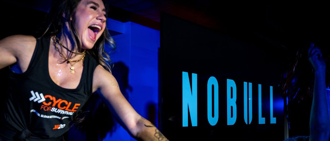 Indoor cycling instructor wearing a Cycle for Survival tank top during a high‑energy ride, with NOBULL branding in a dark studio setting.