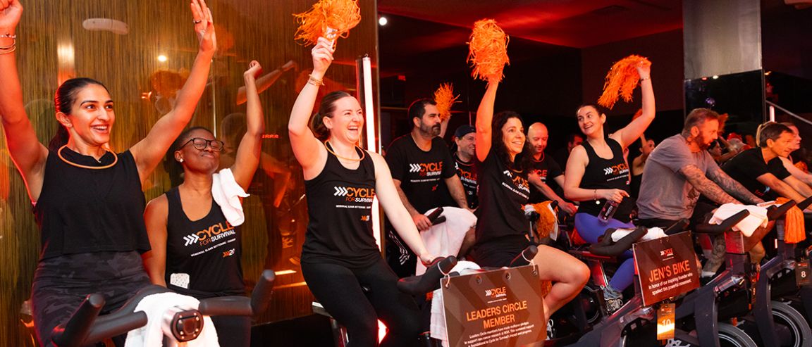 People wearing shirts that read “Cycle for Survival” cheer with orange pom-poms at a Cycle for Survival event. 
