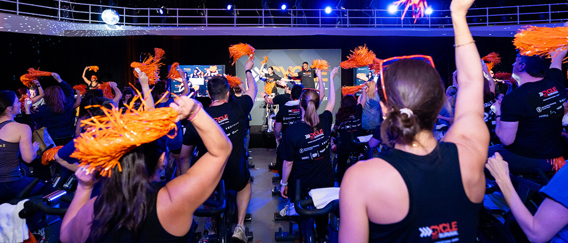 Cycle for Survival participants ride stationary bikes and wave orange pom-poms during a large indoor fundraising event. 