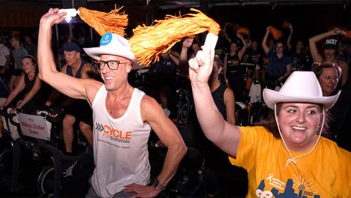 Two fitness instructors at a rare cancer fundraiser stand on a stage in front of a backdrop that says “Cycle for Survival.”