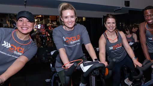 Four smiling people on exercise bikes at a Cycle for Survival event.