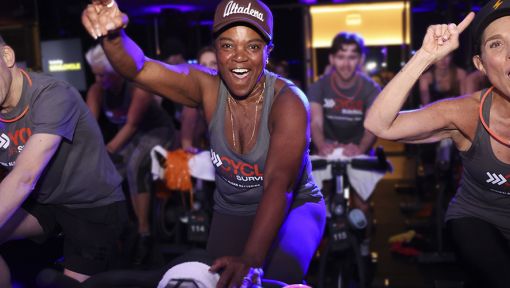 Participants in matching Cycle for Survival shirts ride stationary bikes energetically in a dimly lit indoor cycling event.