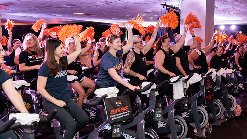 Cycle for Survival riders cheer and wave orange pom poms during a high-energy indoor team cycling event.