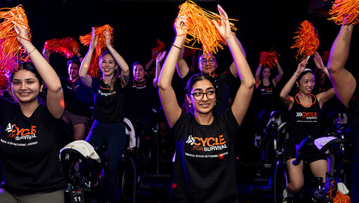 Group of riders on stationary bikes raise orange pom-poms in a dark studio, wearing Cycle for Survival shirts.