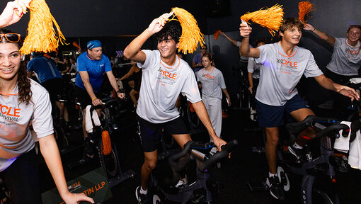 Participants ride stationary bikes and wave orange pom-poms at a Cycle for Survival fundraising event.