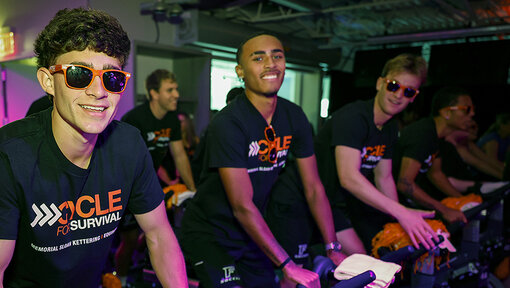 People ride stationary bikes in a neon-lit studio, wearing matching Cycle for Survival shirts and sunglasses.