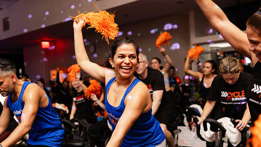 Cycle for Survival participants ride stationary bikes and wave orange pom-poms during a high-energy indoor event.