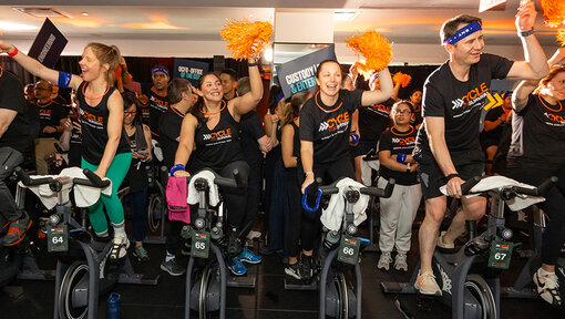 Participants ride stationary bikes in a crowded space, raising orange pom-poms during a Cycle for Survival event.