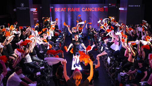 Large group of participants on stationary bikes raise orange pom-poms and white towels beneath a “Beat Rare Cancers” sign.