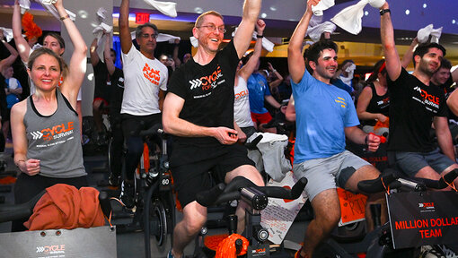 Group of participants on stationary bikes lift white towels in a bright Cycle for Survival studio during a high-energy ride.