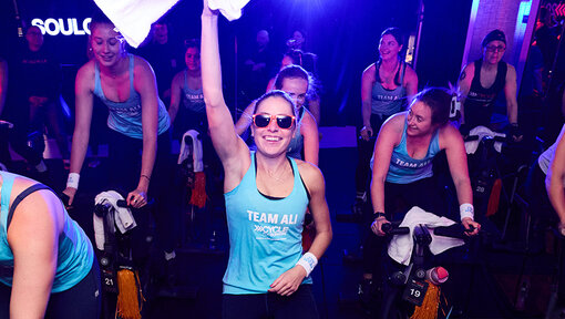 Participants ride stationary bikes under purple lighting, with one rider in front raising a white towel during a Cycle for Survival event.