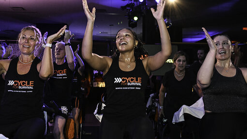 Participants on stationary bikes clap overhead in a dim studio, wearing Cycle for Survival shirts during a group ride.