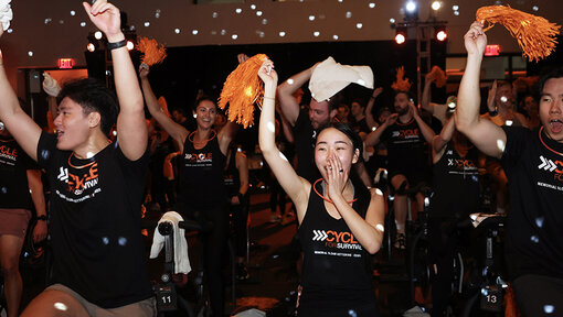 People ride stationary bikes in a dim studio, raising orange pom-poms and towels during a Cycle for Survival group ride.