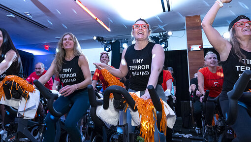 Cycle for Survival participants ride stationary bikes and wave orange pom-poms during an indoor event with bright lighting.