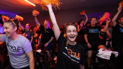 Cycle for Survival participants ride stationary bikes and raise orange pom-poms during a high-energy indoor event.