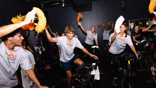 Cycle for Survival participants ride stationary bikes and wave orange pom-poms during a lively indoor fundraising event.