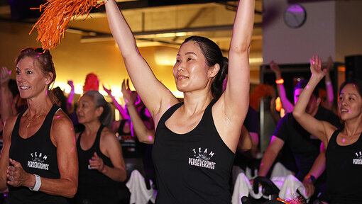 Cycle for Survival participants celebrate and ride together during an indoor event, raising arms beneath colorful lights.