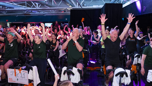 Cycle for Survival participants ride stationary bikes and raise their arms during a high-energy indoor event with colorful lights.