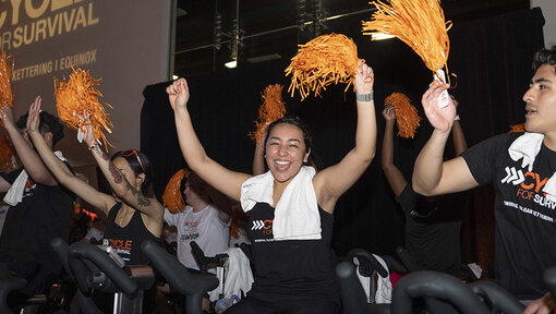 Cycle for Survival participants ride stationary bikes and wave orange pom-poms during a celebratory indoor event.