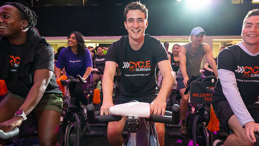 Cycle for Survival participants ride stationary bikes side by side during an indoor event under bright stage lighting.