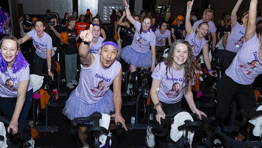 Cycle for Survival participants ride stationary bikes and wave pom-poms during a spirited indoor event with colorful lighting.