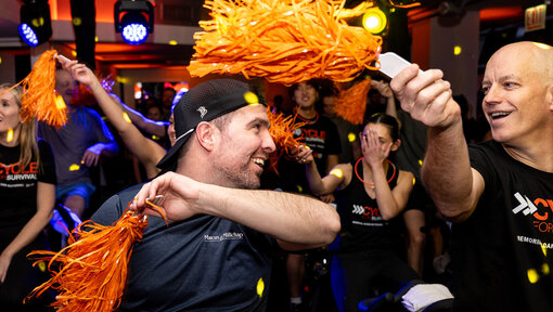 A group cheers with orange pom-poms at a Cycle for Survival event as riders and supporters celebrate together.