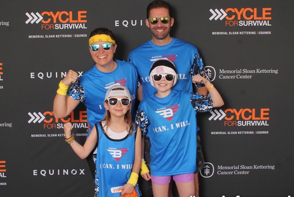 A family of four stands in front of a Cycle for Survival banner, posing for a photo. All are wearing blue shirts that say “I can. I will.” 