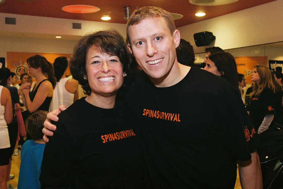 A man and woman stand shoulder to shoulder in a crowded room. Both are smiling and wearing black T-shirts that say “SPIN4SURVIVAL.” 
