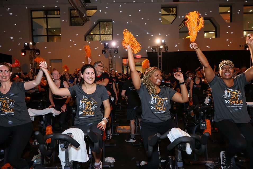 Four Amazon Web Services employees ride stationary bikes, cheer with orange pom-poms, and celebrate at an indoor Cycle for Survival event.