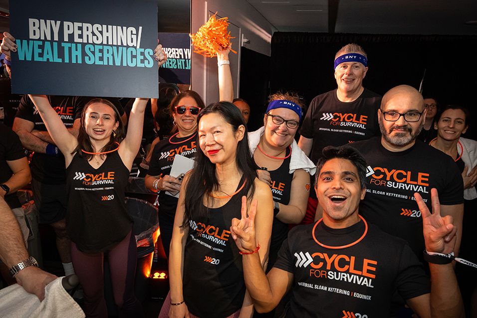 BNY employees wearing Cycle for Survival T-shirts celebrate at a Cycle for Survival event. One holds a sign reading “BNY PERSHING/WEALTH SERVICES.”