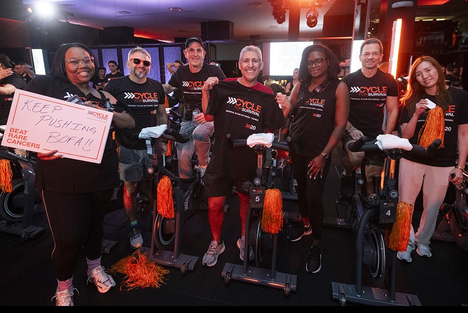 Seven Bank of America employees wearing Cycle for Survival shirts gather at a Cycle for Survival event. One holds a sign reading “KEEP PUSHING BOFA.”