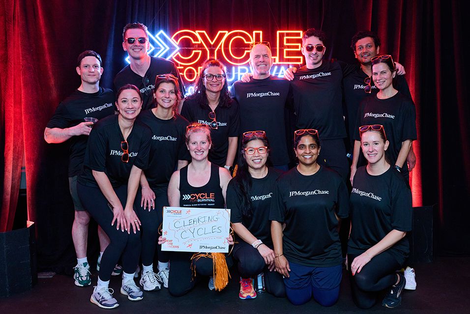 JPMorganChase employees, wearing shirts reading “JPMorganChase,” gather at a Cycle for Survival event. One rider holds a sign reading “Clearing Cycles.”