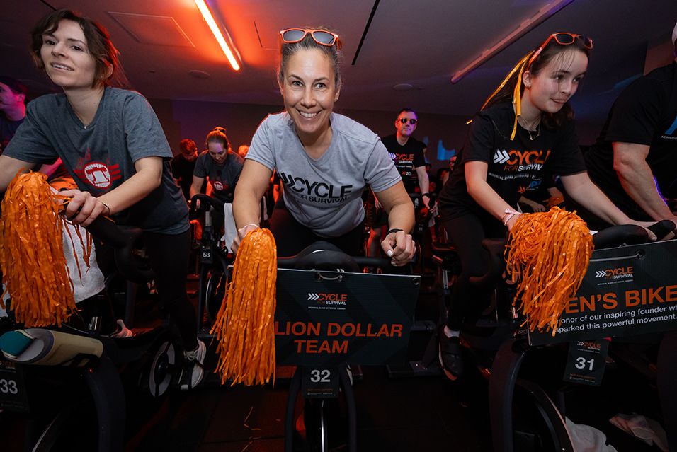 Gia Fruscione cycles indoors with fellow Cycle for Survival riders, many holding orange pom-poms, wearing a gray event shirt. 